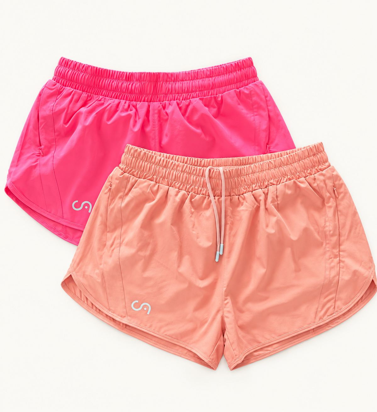 Two pairs of athletic shorts in bright pink and peach-orange, arranged overlapping on a clean white background. Each pair has an elastic waistband, a drawstring, and a small white stylized logo near the lower left leg.